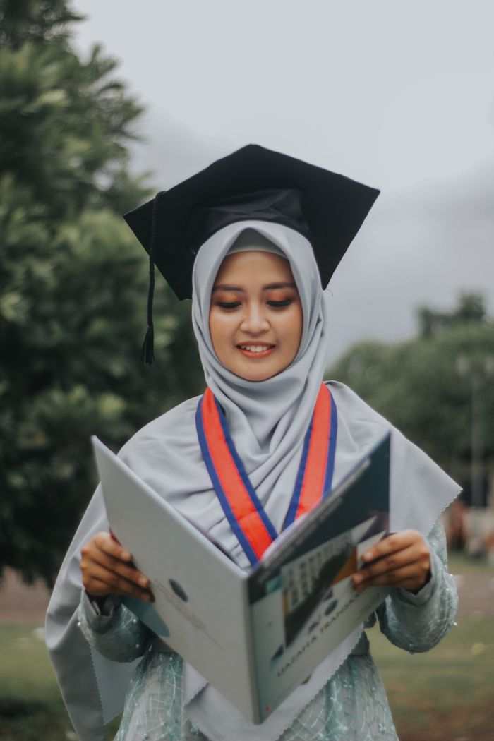 Young graduate in hijab and gown holding diploma, celebrating education success outdoors.