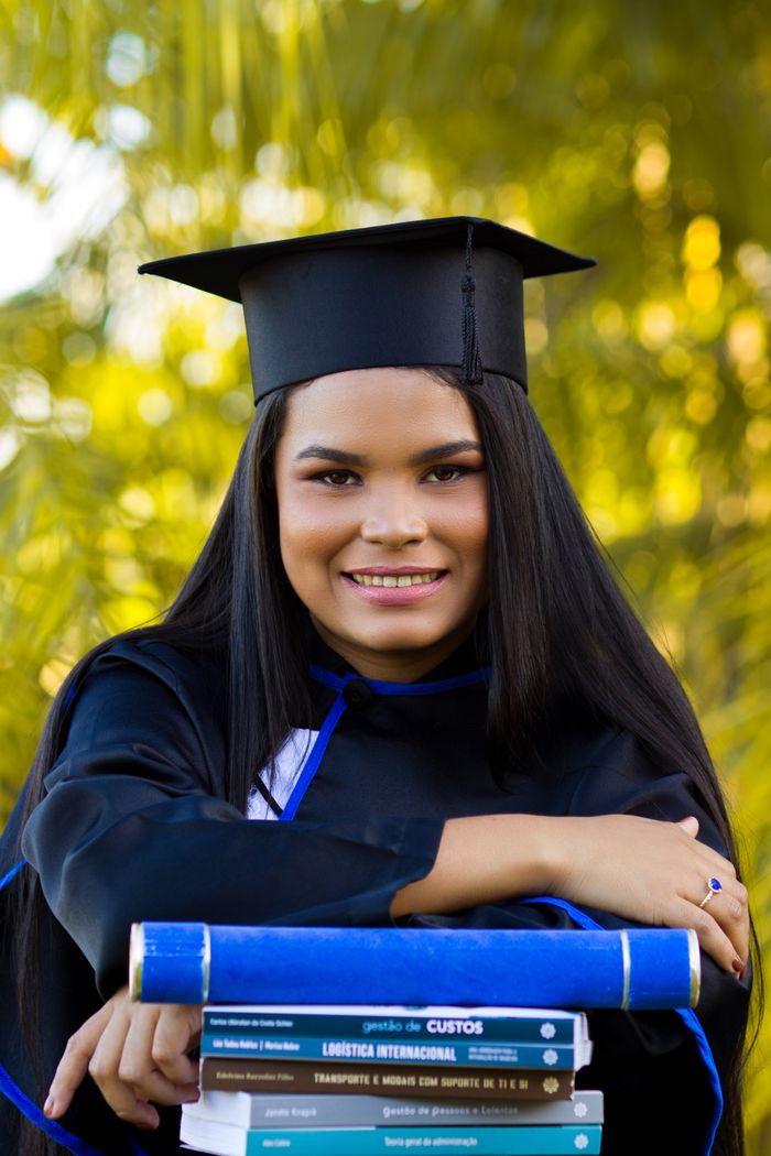 Glad young ethnic woman in graduation cap with textbooks smiling and looking at camera on blurred background of greenery